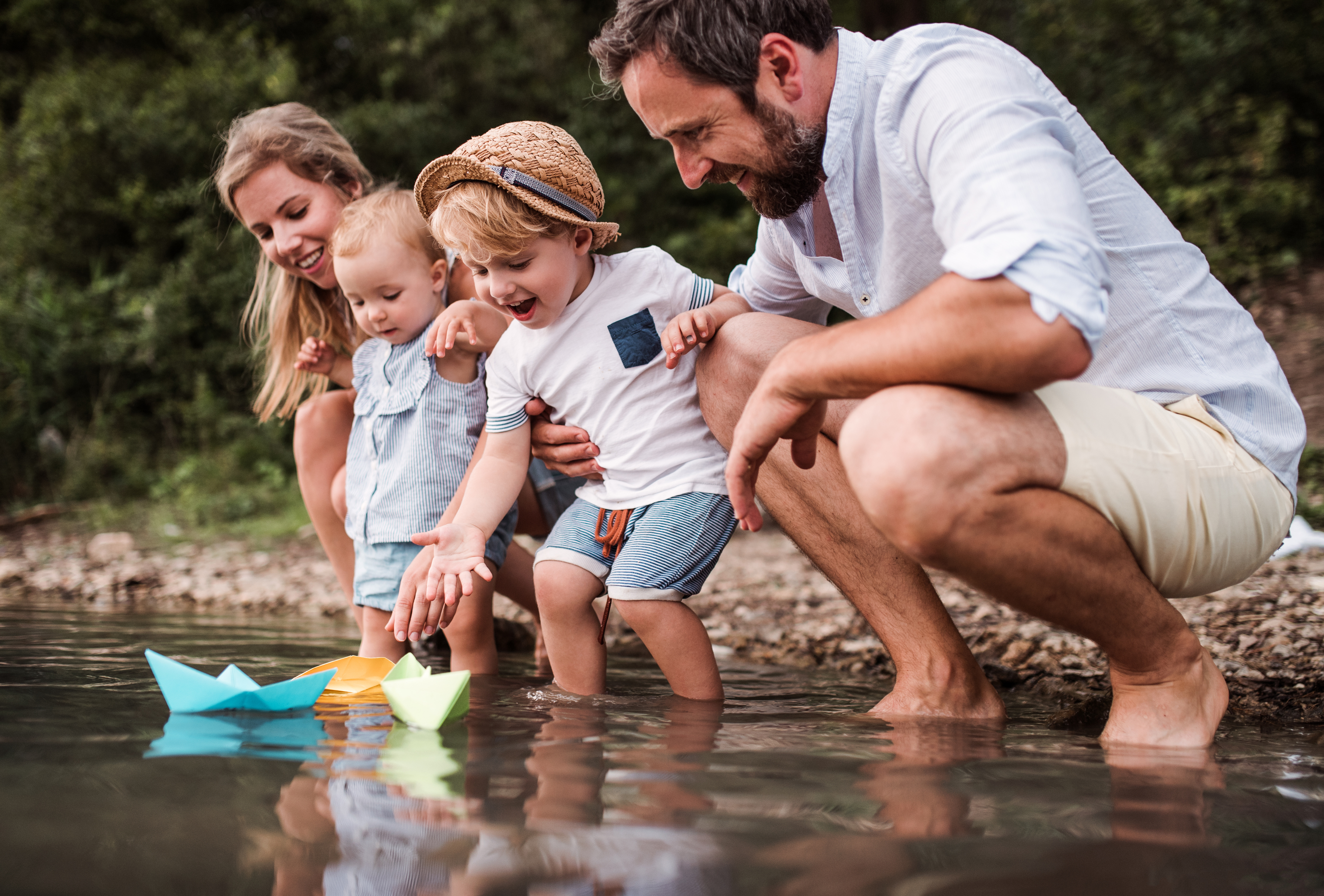 Parents with two young children float paper boats in a river.